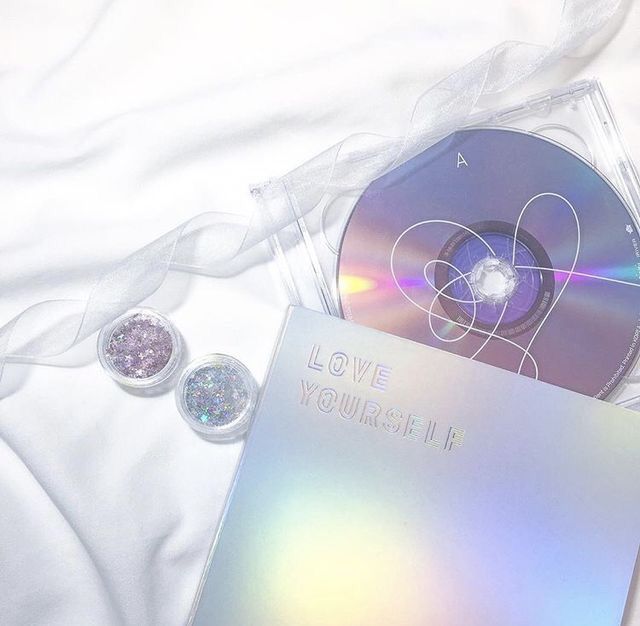 iridescent purple and blue cd and album cover laid on a white cotton bed sheet. there are two pots of glitter on the left - one purple and one blue